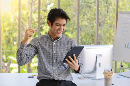 Portrait of smart young asian businessman holding tablet and feeling glad and raised fist in work room with computer and meeting board and accesories on desk.の写真素材