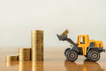 Business, Money,  Finance and Saving  Concept. Close up of miniature mini loader truck contain 4 coins and puting to top of stack of gold coins on wooden table with copy space.の写真素材