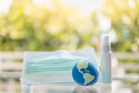 Global Healthcare, Coronavirus, Covid-19 Protection Concept. Close up of mini world ball with surgical face mask and mini bottle alcohol spray with green nature background.の写真素材