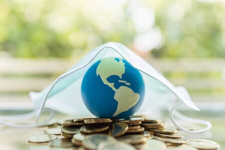 Global Money, Business, Healthcare in Cornavirus (COVID-19) Situation Concept. Mini world ball on pile of gold coins under surgical face mask with copy space.の写真素材