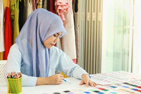 Portrait of beautiful asian muslim woman designer with hijab drawing design with coloring pencil in her home working studio room with sample color and pattern cloth, dress, jacket and material.の写真素材