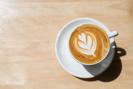 Top view of white cup of hot coffee latte with milk foam heart shape art on wooden table under morning sunlight and shadow.の写真素材