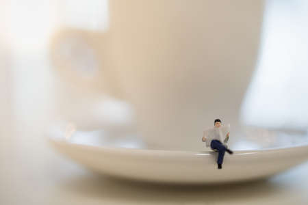 Closeup of businessman miniature figure people sitting and reading a newspaper on white plate and cup of hot coffee.の写真素材