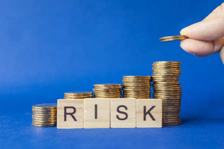 Business, Money and Risk Management Concept. Closeup of man hand holding two gold coin and put to top of stack of coins with English letter wooden block on blue background.の写真素材