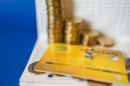 Business, E-Commerce and Money Concept. Closeup of miniature shopping cart or trolley on mockup fake yellow credit card with pile and stack of gold coin on bank passbook with blue background.の写真素材