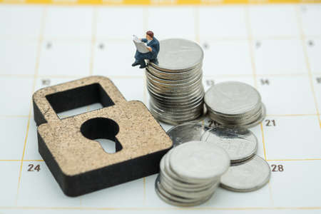 Money , Planning and Security Concept. Businessman miniature figure people sitting and reading a newspaper on stack of coins with wooden key lock symbol on calendar.の写真素材