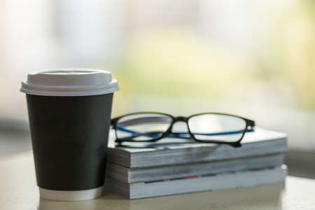 Closeup of takeaway papercup of hot coffee with stack of books and reading glasses on wooden table.の写真素材
