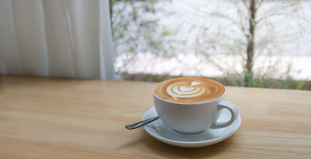 Closeup of white cup of hot coffee latte with milk foam art on wooden table.
