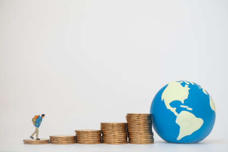 Global Business Travel and Saving Concept. Traveler miniature with backpack walking on stack of gold coins with mini world ball on white background.の写真素材