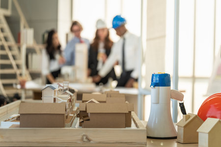 Closeup of wireless loudspeaker megaphone  with orange hard safety helmet hat and wooden mini building house model with businesssman, busineeewoman and engineer as background.の写真素材