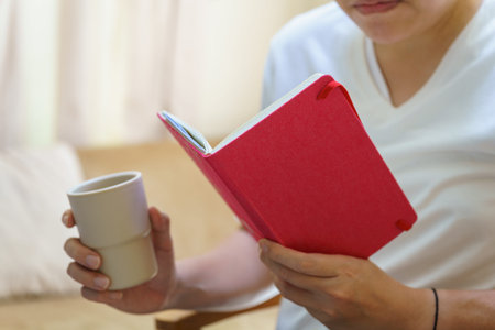 A man wearing white t-shirt sitting and reading a book and holding a plastic cup of hot coffee.