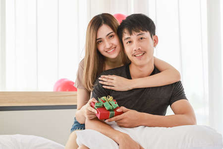 Couple Love and Valentine's Day Concept. Young smiling Asian man and woman looking to camera with beautiful giving gift box on bed in bedroom with red balloon.の写真素材