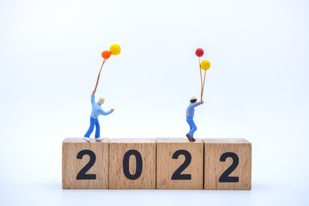 2022 New Year and Family Concept. Closeup of two kid miniature figures with balloon plaing on stack of wooden number block on white background.の写真素材