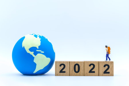 2022 New Year and Travel Concept. Closeup of woman traveler miniature figures with backpack standing on stack of wooden number block with mini world ball on white background.の写真素材