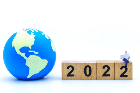 2022 New Year and Business Concept. Closeup of businessman miniature figures sitting and reading a book or newspaper on wooden number block with mini world ball on white background.の写真素材