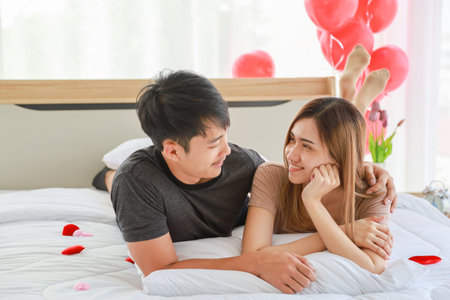 Couple and Valentine Day Concept. Asian smiling female and male on white blanket on bed in bedroom with mini red heart shape pillow.の写真素材