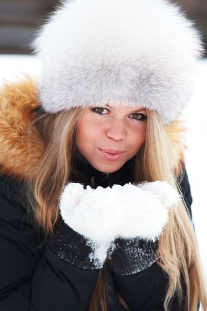 Young woman in a fur-cap in a winter forest blowing snowflakesの写真素材