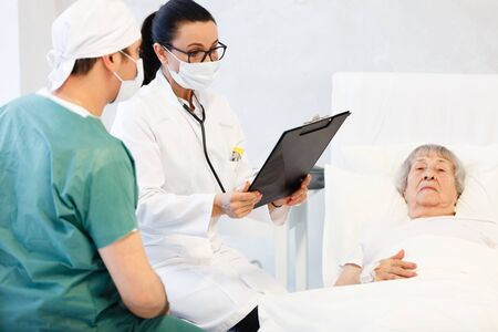 healthcare in hospital. male surgeon and female doctor or nurse making medical examination of old lady lying in bed and getting treatmentの写真素材