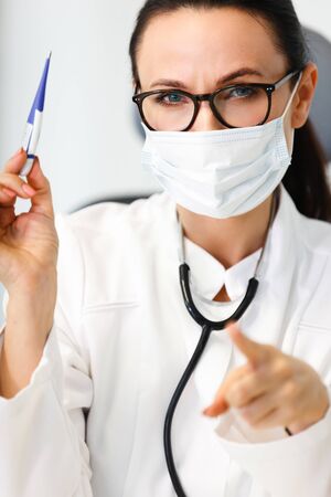 Healthcare in hospital. Female doctor or nurse in medical mask closeup shows electronic thermometer and calling for action to prevent flu, fever and virus infection in medical clinic cabinetの写真素材