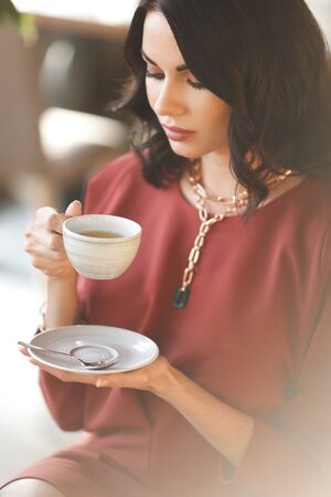 beautiful brunette business lady in modern gems and fashionable burgundy dress having a lunch in cafe. Woman sits by the table and holds cup of teaの写真素材