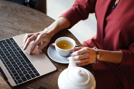 Hands closeup of business lady working on laptop and holding cup of tea in cafe while having lunch. Woman sits by the table with portable computerの写真素材