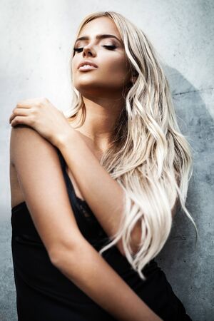 Portrait of blond young woman with beautiful makeup and hair in a classic black dress standing near grey wall. Fashion model posing in elegant clothes outdoorsの写真素材