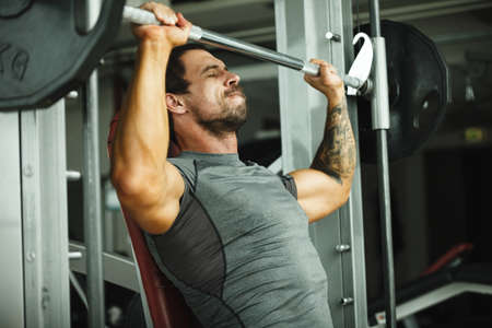 Fitness in gym, sport and healthy lifestyle concept. Handsome athletic man in grey shirt making exercises. Bodybuilder male model training chest muscles with barbell.の写真素材