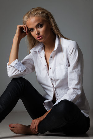 Portrait of blond young woman with beautiful makeup and hairstyle in white shirt sits on gray...