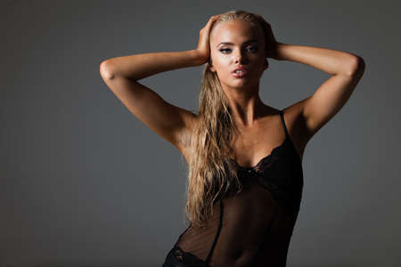 Portrait of blond young woman with beautiful makeup and hairstyle in a black lace bodysuit stands on gray background. Fashion model poses in elegant clothes in studioの写真素材