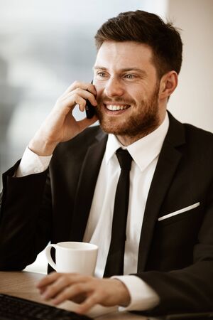 Business concept. Young businessman sitting at the office table happy talking on a cell phone getting good news about his work. Man in suit indoors on glass window backgroundの写真素材
