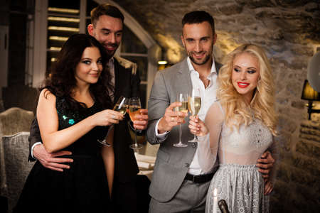 Party, holiday with friends concept. Four people with champagne glasses celebrating and toasting in restaurant. Two men and two women in elegant evening clothes, suits and dresses night out indoorsの写真素材