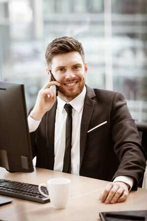 Business concept. Young businessman sitting at the office table happy talking on a cell phone getting good news about his work. Man in suit indoors on glass window backgroundの写真素材