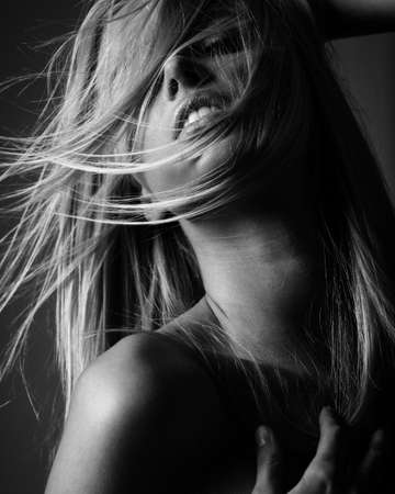 Portrait of blond young woman with beautiful makeup and flying wild hair in black lace dress stands on gray background. Fashion model poses in elegant clothes in studio. Black and white, monochromeの写真素材