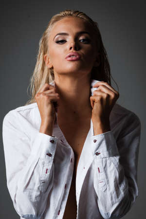 Portrait of blond young woman with beautiful makeup and hairstyle in white shirt stands on gray background. Fashion model poses in elegant clothes in studioの写真素材