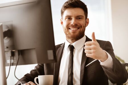 Business concept. Successful young businessman at work. Manager sitting at the office table happy showing thumb up. Man smiling in suit indoors on glass window backgroundの写真素材
