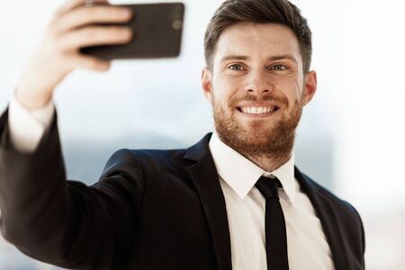 Business concept. Happy smiling young businessman at the office standing and making selfie or video chat conference using cell phone at work. Man in suit indoors on glass window backgroundの写真素材