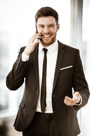 Business concept. Happy smiling young businessman standing in office talking on a cell phone getting good news about his work. Man in suit indoors on glass window backgroundの写真素材