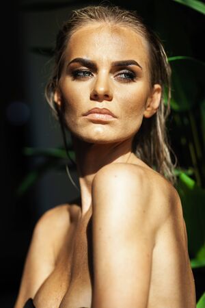 Relaxation and spa concept. Closeup portrait of a sexy fashionable lady in a black swimsuit. Woman with wet hair and makeup poses in green plants in swimming pool in natural sunlightの写真素材