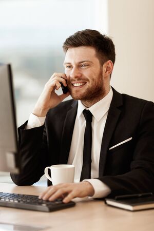 Business concept. Young businessman sitting at the office table happy talking on a cell phone getting good news about his work. Man in suit indoors on glass window backgroundの写真素材