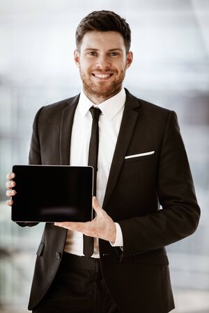 Business concept. Happy smiling young businessman standing in office demonstrating wireless touchpad tablet computer. Man in suit indoors using internet device on glass window backgroundの写真素材
