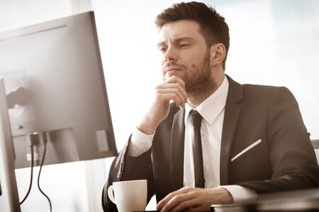 Business crisis concept. Young businessman sitting at the office table busy talking on a cell phone resolving a very serious work problem. Man in suit indoors on glass window backgroundの写真素材
