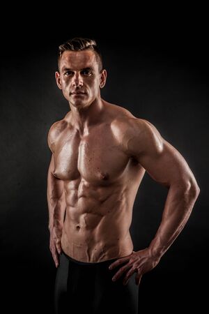 Fitness in gym, sport and healthy lifestyle concept. Handsome athletic man showing his trained body on black background. Bodybuilder male model training biceps muscles with dumbbell.の写真素材