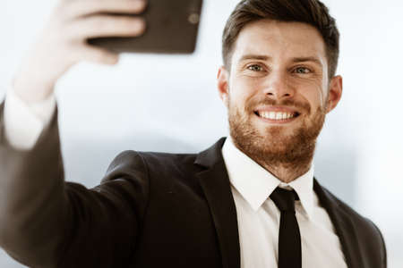 Business concept. Happy smiling young businessman at the office standing and making selfie or video chat conference using cell phone at work. Man in suit indoors on glass window backgroundの写真素材