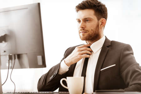 Business concept. Successful young businessman at work. Manager sitting at the office table, working on computer and straightens a tie. Busy man in suit indoors on glass window backgroundの写真素材