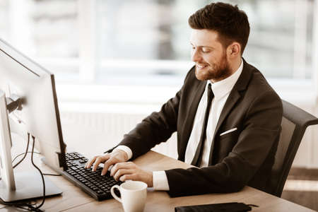 Business succsess concept. Happy smiling young businessman sitting in office and working on computer. Man in suit indoors on glass window backgroundの写真素材
