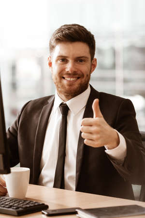 Business concept. Successful young businessman at work. Manager sitting at the office table happy showing thumb up. Man smiling in suit indoors on glass window backgroundの写真素材