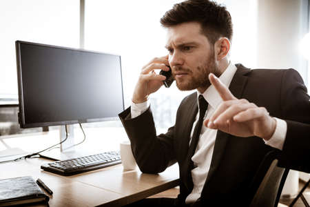 Business crisis concept. Young businessman sitting at the office table busy talking on a cell phone resolving a very serious work problem. Man in suit indoors on glass window backgroundの写真素材