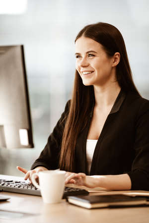 Business succsess concept. Happy smiling young businesswoman in office gets good news while work on computer. Woman in suit indoors on glass window backgroundの写真素材