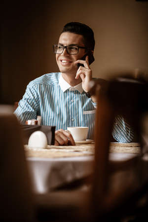 LGBTQ community lifestyle concept. Young homosexual man sits at the table in old-fashioned city cafe. Handsome gay male businessman talks on a phone while having a break at lunchの写真素材