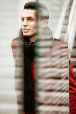 LGBTQ community lifestyle concept. Young homosexual man stands behind glass. Handsome fashionable gay male model poses in cityscape outdoors. Wears red coat, gloves, and black scarfの写真素材